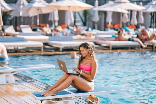 Cute young woman sunbathing sitting in front of the pool, working with a laptop at summer day
