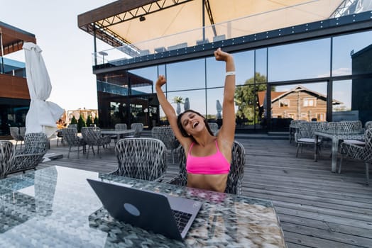Cute young woman with crazy emotions sitting on terrace, while using laptop for work at summer day