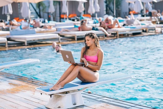 Cute young woman sunbathing sitting in front of the pool, working with a laptop at summer day
