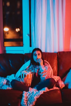 Beautiful young girl eating popcorn watching movie at home in a dark room with a popcorn bucket and playing with her dog and sits in front of a monitor or TV watching movie. Crazy emotions. High quality photo