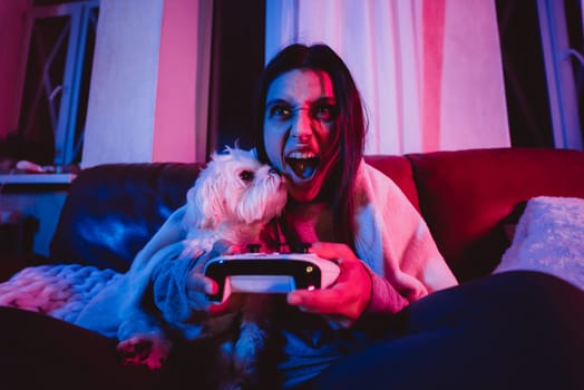 Close up portrait of A gamer or a streamer girl at home in a dark room with a game controller playing with her dog and sits in front of a monitor or TV. Crazy emotions. High quality photo