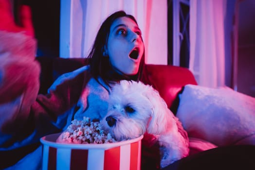 Close up portrait of Beautiful young girl at home in a dark room with a popcorn bucket playing with her dog and sits in front of a monitor or TV watching movie. Crazy emotions. High quality photo