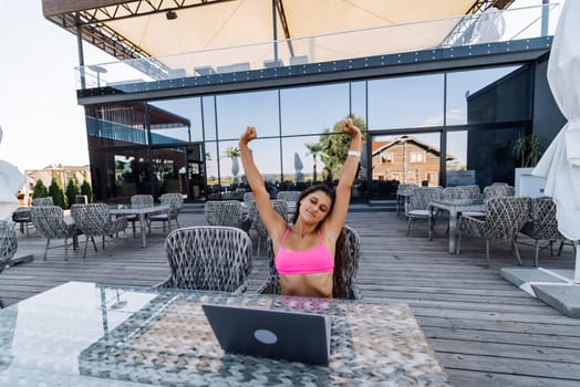 Cute young woman with crazy emotions sitting on terrace, while using laptop for work at summer day