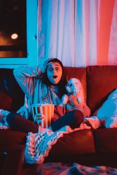 Beautiful young girl watching movie at home in a dark room with a popcorn bucket and playing with her dog and sits in front of a monitor or TV watching movie. Crazy emotions. High quality photo