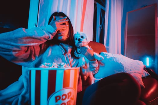 Close up portrait of Beautiful young girl hide her eyes watching movie at home in a dark room with a popcorn bucket playing with her dog and sits in front of a monitor or TV watching movie. Crazy emotions. High quality photo
