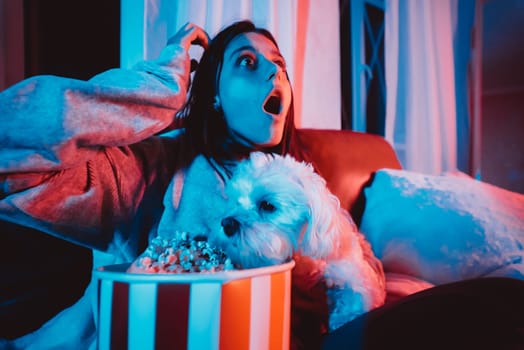 Close up portrait of Beautiful young girl at home in a dark room with a popcorn bucket playing with her dog and sits in front of a monitor or TV watching movie. Crazy emotions. High quality photo