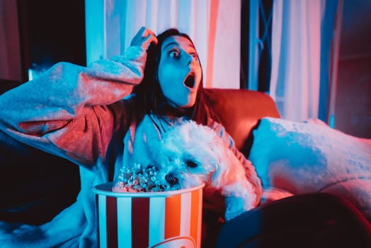 Close up portrait of Beautiful young girl at home in a dark room with a popcorn bucket playing with her dog and sits in front of a monitor or TV watching movie. Crazy emotions. High quality photo