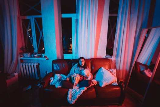 Beautiful young girl watching movie at home in a dark room with a popcorn bucket playing with her dog and sits in front of a monitor or TV watching movie. Crazy emotions. High quality photo