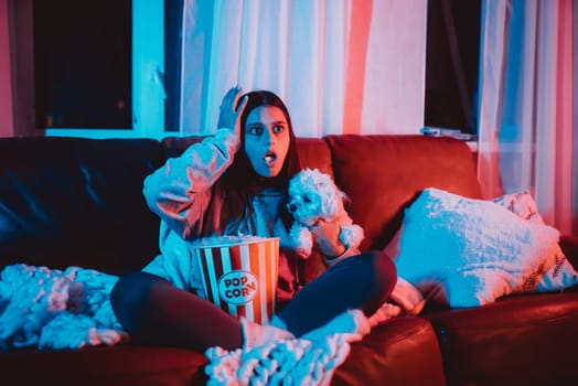 Beautiful young girl watching movie at home in a dark room with a popcorn bucket playing with her dog and sits in front of a monitor or TV watching movie. Crazy emotions. High quality photo