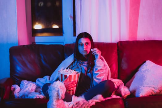Beautiful young girl eating popcorn watching movie at home in a dark room with a popcorn bucket and playing with her dog and sits in front of a monitor or TV watching movie. Crazy emotions. High quality photo