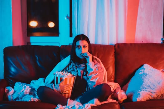 Beautiful young girl eating popcorn watching movie at home in a dark room with a popcorn bucket and playing with her dog and sits in front of a monitor or TV watching movie. Crazy emotions. High quality photo