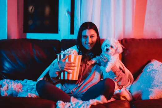 Beautiful young girl smiles watching movie at home in a dark room with a popcorn bucket and playing with her dog and sits in front of a monitor or TV watching movie. Crazy emotions. High quality photo