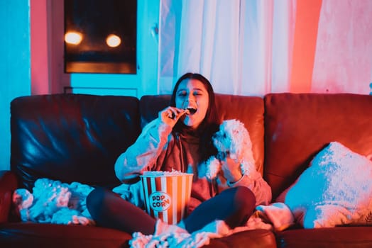 Beautiful young girl eating popcorn watching movie at home in a dark room with a popcorn bucket and playing with her dog and sits in front of a monitor or TV watching movie. Crazy emotions. High quality photo