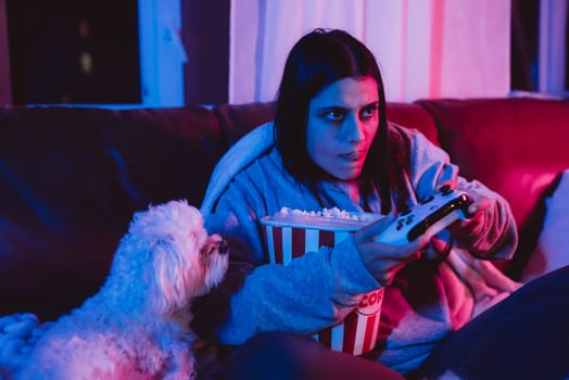 A gamer or a streamer girl at home in a dark room with a game controller and popcorn bucket playing with her dog and sits in front of a monitor or TV. High quality photo