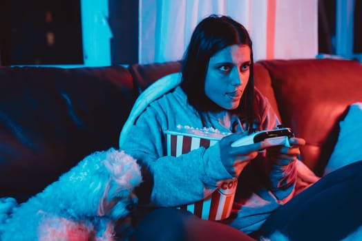 A gamer or a streamer girl at home in a dark room with a game controller and popcorn bucket playing with her dog and sits in front of a monitor or TV. High quality photo