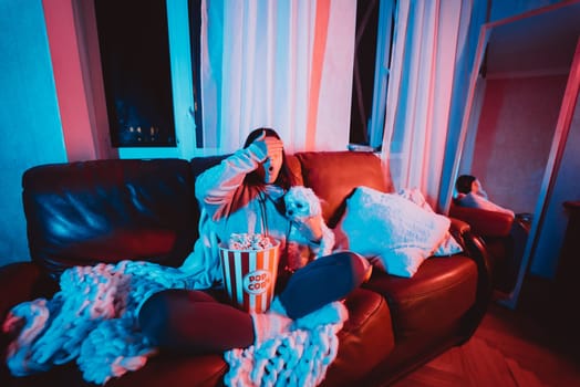 Close up portrait of Beautiful young girl hide her eyes watching movie at home in a dark room with a popcorn bucket playing with her dog and sits in front of a monitor or TV watching movie. Crazy emotions. High quality photo