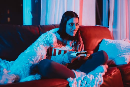 A gamer or a streamer girl at home in a dark room with a game controller and popcorn bucket playing with her dog and sits in front of a monitor or TV. High quality photo