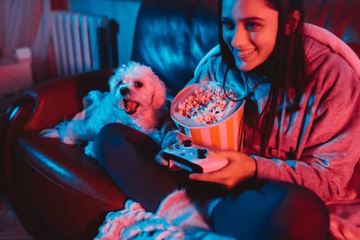 A gamer or a streamer girl at home in a dark room with a game controller and popcorn bucket playing with her dog and sits in front of a monitor or TV. High quality photo