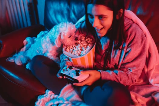 A gamer or a streamer girl at home in a dark room with a game controller and popcorn bucket playing with her dog and sits in front of a monitor or TV. High quality photo