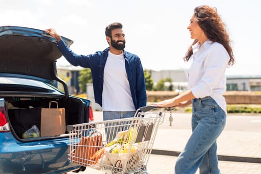 Happy arab man opening car trunk for packing products, woman standing with shopping cart full of fresh food after successful shopping in supermarket