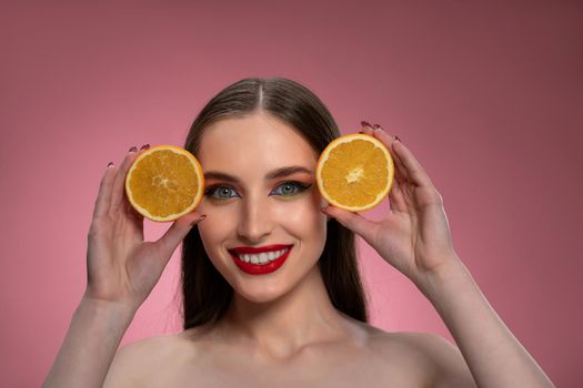 Portrait of a positive young woman with oranges sliced in her hands looking charming. Charming joyful funny lady with long hair isolated on pink background.