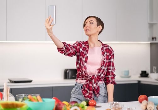 Pretty woman happy taking selfie using her smartphone at her new home while cooking fresh salad wearing a plaid shirt with a bob hair style. Healthy food leaving - vegan concept. 