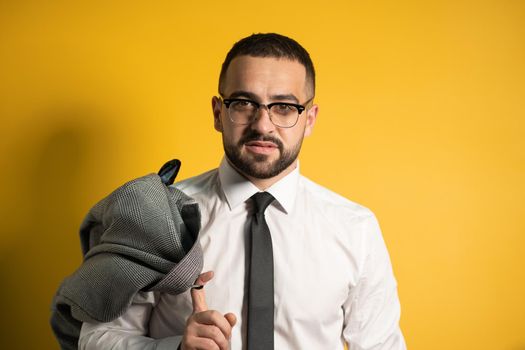 Looking serious business man with trendy beard dressed in greyish suite posing holding his jacket on his shoulder hanging it behind looking isolated on yellow background.