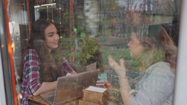 View through glass two student girls talking while enjoying coffee together in a coffee shop with customers walking on the background as they sit at a table chatting working on laptop. 4K footage.