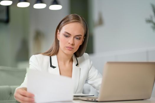 Portrait of a beautiful businesswoman in white jacket working with documents in her hands sitting in front of laptop. Office worker looking at her working place.