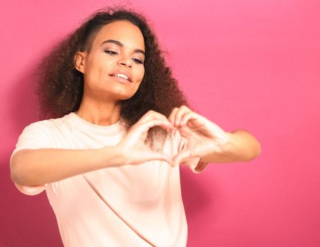 Sending love with gesture of a tender looking beautiful young African American woman looking positively at camera wearing peachy t-shirt isolated on pink background. Beauty concept. 