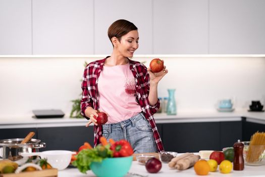 Housewife cooking apple pie standing at the kitchen wearing plaid shirt with a short hair. Holding an apple in hand happy woman dancing at kitchen with a sexy eye contact on camera. 