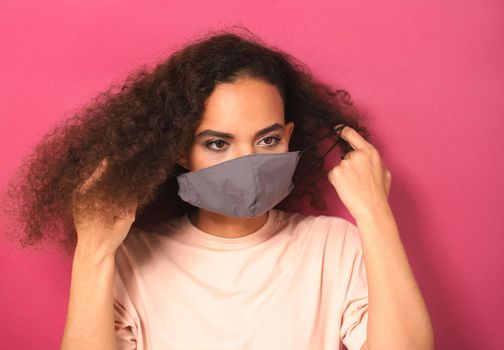 Putting on reusable face mask African American charming girl in peachy t-shirt, to prevent others from corona COVID-19 and SARS cov 2 infection isolated on pink background. 