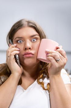 Tired of stress or shocked by a weight loss plan overweight young woman talking on the phone holding a cup with cold water touching chick. Stylish chubby student girl talking on mobile. 