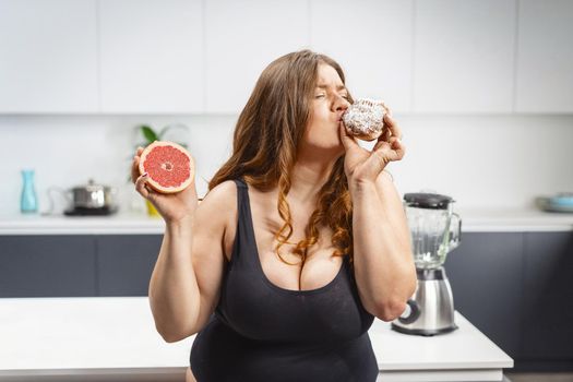 Young fat woman choosing what to eat holding a cake and grapefruit. Beautiful chubby young woman eating unhealthy food. Fat girl comparing between eating cake and fruit. 