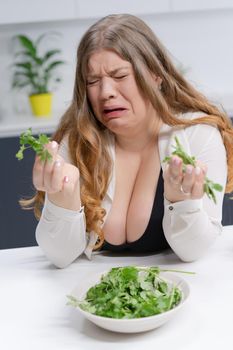 Fat girl not happy holding fresh salad casting. Curvy body young woman with long blond hair sitting on modern kitchen. Dieting and nutrition concept. 