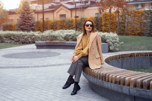 Beautiful young autumn french woman in beige coat and sunglasses sits on a flowerbed bench waiting for her date or girlfriends. Soft warm toned photo. 