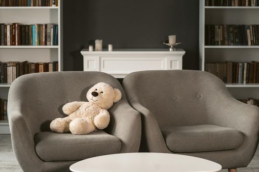 Teddy sitting on armchair. Teddy bear on grey chair next to empty one in interior. Beautiful living room with teddy bear on chair.