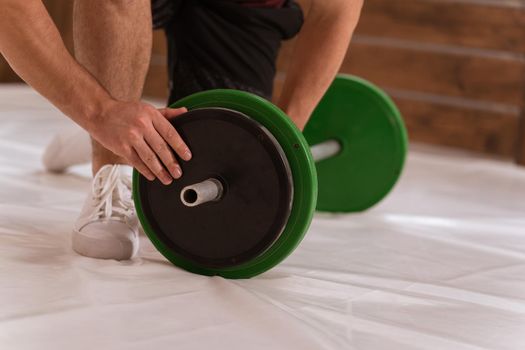 Fixing barbell young man bow on a knee adding a black and green plates, equipment for weight training concept. Sports equipment for training. Weight loss, healthy lifestyle concept.