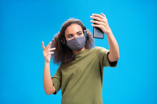 Talking on video call young African American girl using her smartphone with headphones on in olive t-shirt, reusable face mask, to prevent others from corona COVID-19 isolated on blue background. 