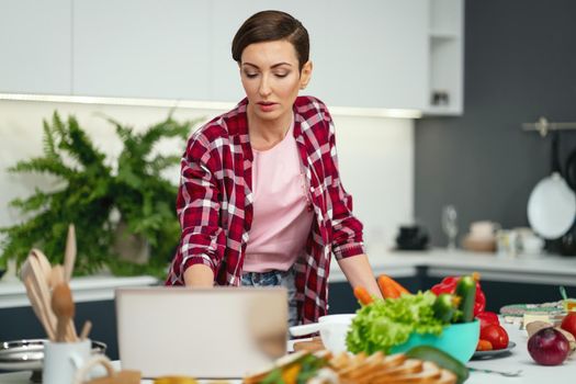 Making a video call using laptop computer housewife showing her new house and new kitchen while cooking or baking holding eggs in a hand. Young woman cooking in the kitchen. New house concept.