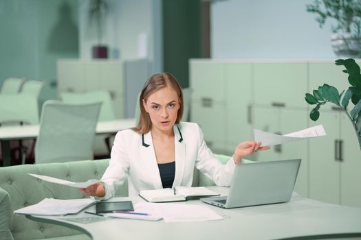 Dissatisfied or tired blond businesswoman working with documents sitting in front of laptop wearing white official suit. Office worker looking at camera sitting at her working place.