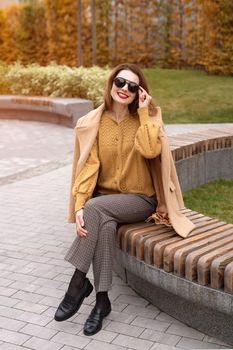 Beautiful young woman in an autumn beige coat and sunglasses sits on a flowerbed bench waiting for her date or girlfriends. Toned photo. 