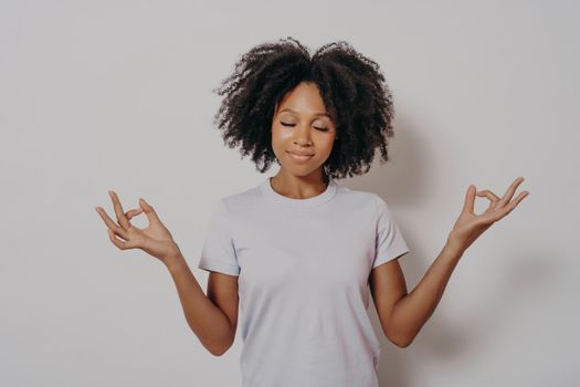 Studio portrait of calm and peaceful young mixed race woman keeping hands in mudra gesture and eyes closed, meditating while standing isolated over white wall. Meditation concept