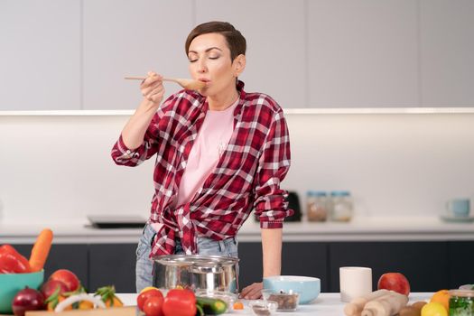 Woman trying a cooked dish using a long wooden spoon. Woman with a short hair cooking a dinner for family standing in the modern kitchen of a new house. Healthy food at home. 