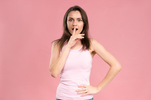 Young caucasian woman with dark hair in trendy pink t-shirt looking at camera with suspicious expression, holding hand on her waist and finger at the lips. Facial expressions, emotions, feelings.