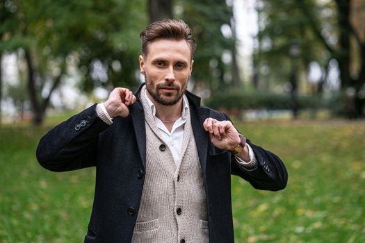 A handsome young man straightens the sides of his dark blue coat. A young business man stands on the street in an autumn coat and cardigan under it looks confidently at the camera.