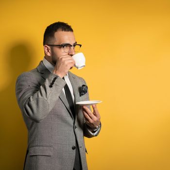 Handsome young bearded business man in eye glasses in formal wear holding a cup of coffee standing sideways isolated on yellow background. 