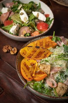 Two bowls of salads with salmon, mozzarella cheese and grilled slices of fruits stands on a table covered with a leather rag. Restaurant concept. Vertical image.