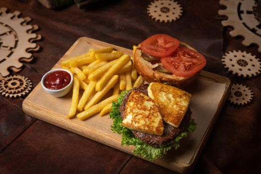 Disassembled beef burger with fried cheese, slices tomatoes served with french fries on a decorative wooden board with tomato sauce with wooden decorative pieces of simple mechanism. 