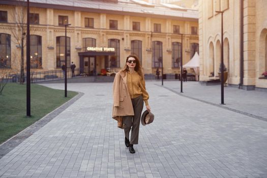 Fashion young woman in sunglasses and hanging on a shoulder autumn beige coat walking on the street happily posing for the camera. Tinted photo. 